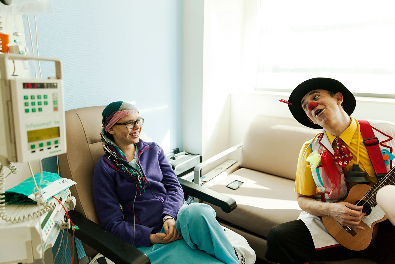 Payaso cantando con guitarra a una chica en el hospital que sonríe y lleva un turbante en la cabeza