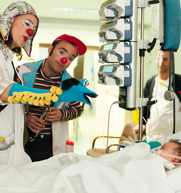 Payaso y payasa animando a un niño tumbado en cama de hospital con marioneta